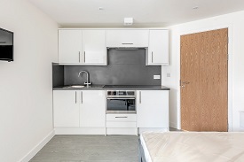 A studio room in 59 West Street. Against the far wall is a kitchen unit with an oven/hob, sink, fridge/freezer and cupboard space. To the right of the kitchen unit is the door to the room. A bed is the right of the door and a television is mounted onto the wall opposite the bed.